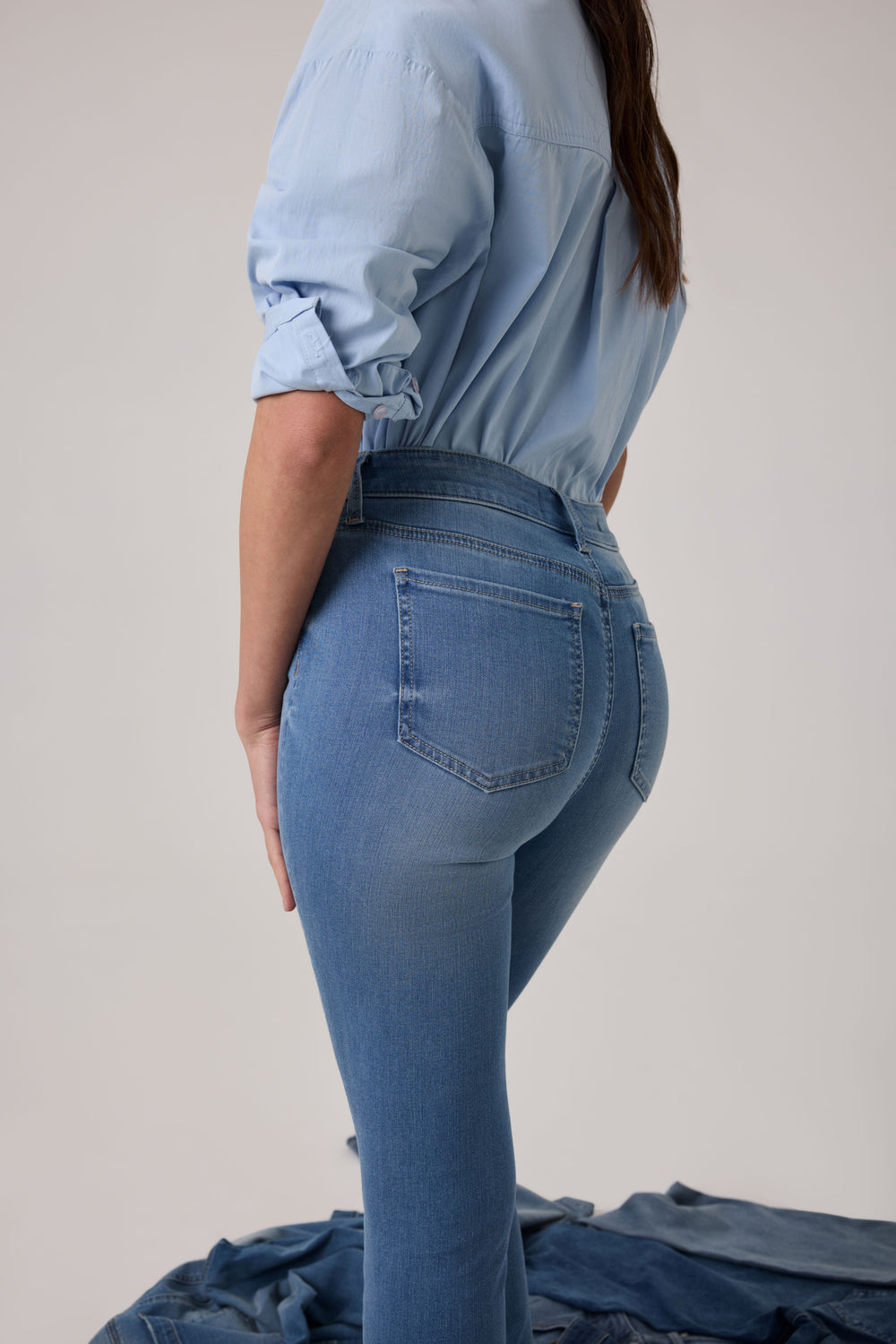A woman wearing a light denim top and medium wash denim jeans with a pile of denim in the background. 